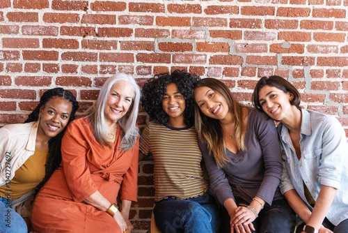 Fotografie Group of five diverse women sitting together, smiling in front of a brick wall