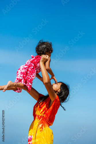 Asian teenage girl in colorful beach dress holding and lifting up her little sister, playing during summer vacation with blue sky on the background