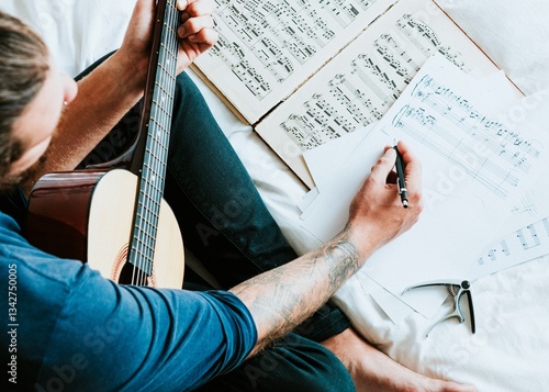Man with guitar writing music. Guitar, music sheets, and pen. Focused on composing music. Creative process with guitar and music sheets. Tattooed arm visible. Musician composing a song on guitar.