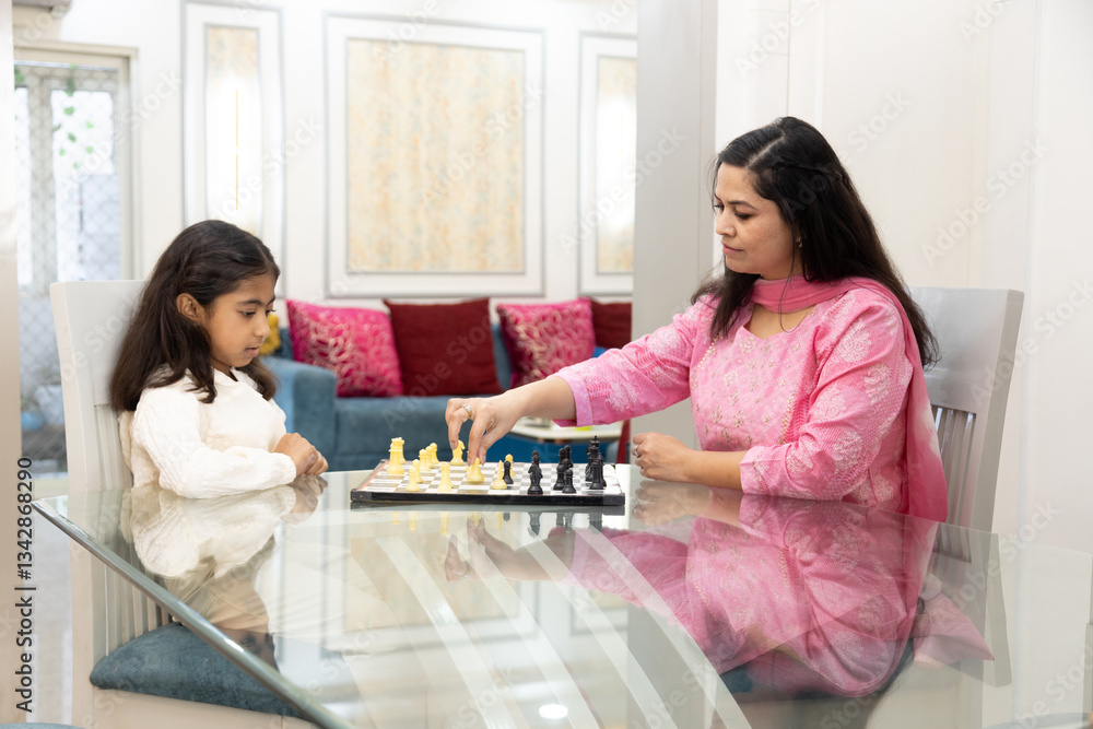 Fototapeta premium Young indian mother and child daughter play chess game at home. Gild kid enjoy Indoor sport with her mum. Challenge and competition. Relationship and bonding.