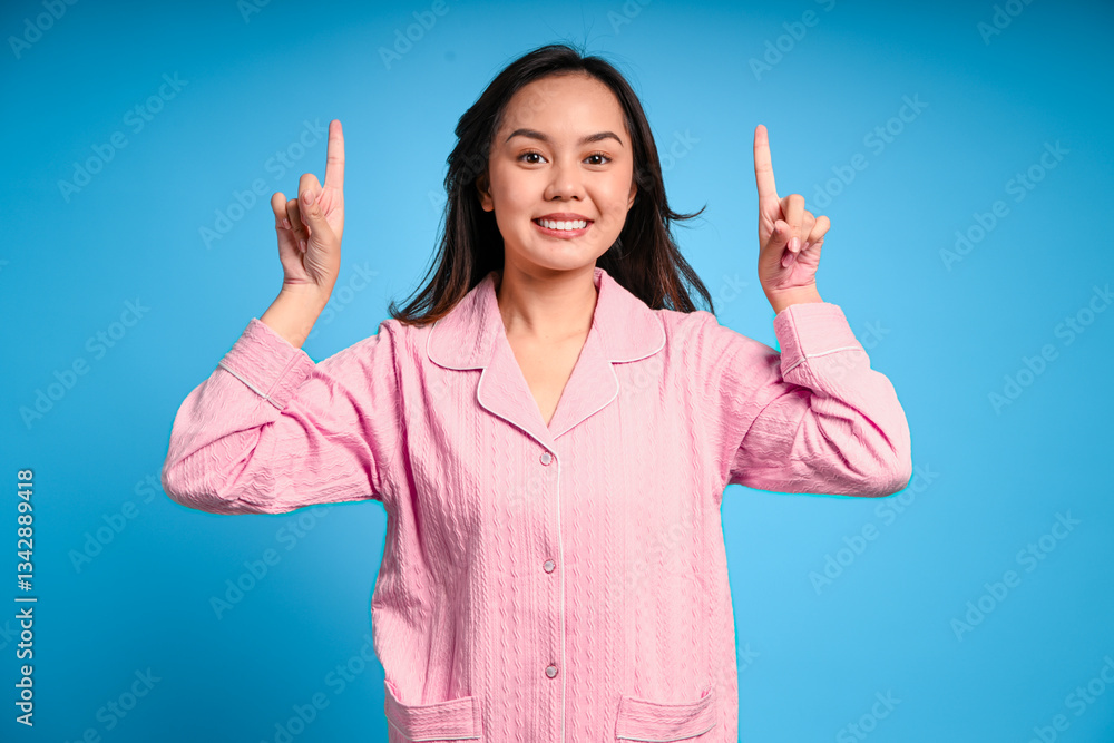 Asian woman in pink pajamas pointing both index fingers upward with an excited expression, standing against a vibrant blue background in a playful and cheerful studio scene Asian woman in pink pajamas pointing both index fingers upward with an excited expression, standing against a vibrant blue background in a playful and cheerful studio scene