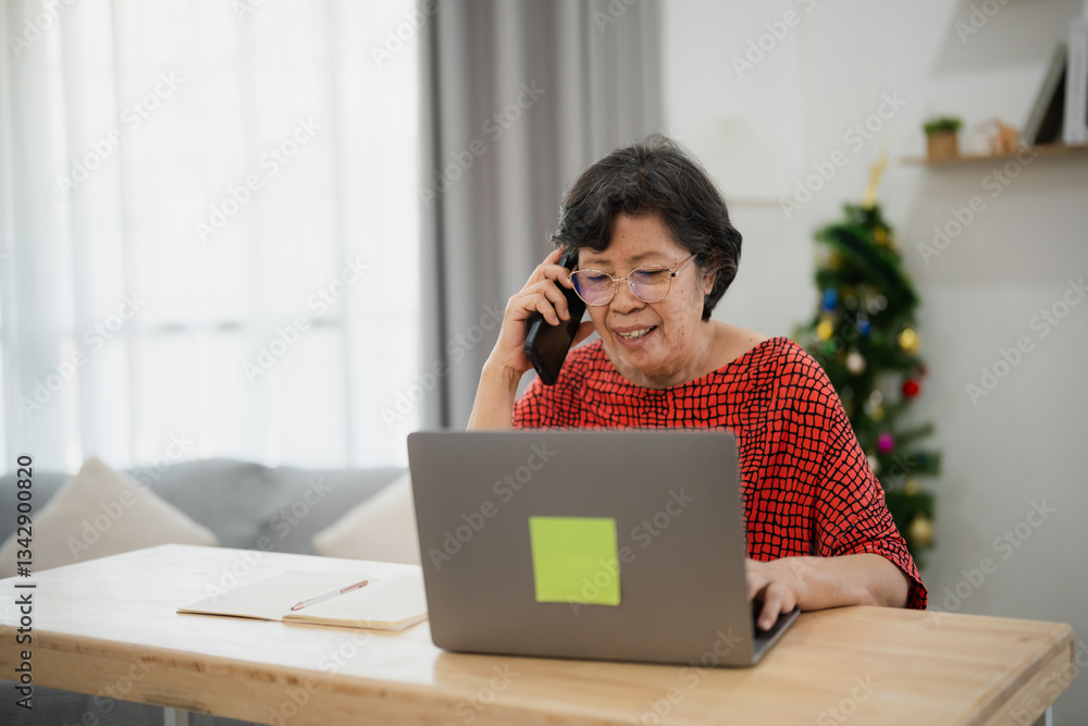 Senior Asian woman using laptop while talking on smartphone at home, enjoying a peaceful moment in the modern living space with holiday decorations Senior Asian woman using laptop while talking on smartphone at home, enjoying a peaceful moment in the modern living space with holiday decorations