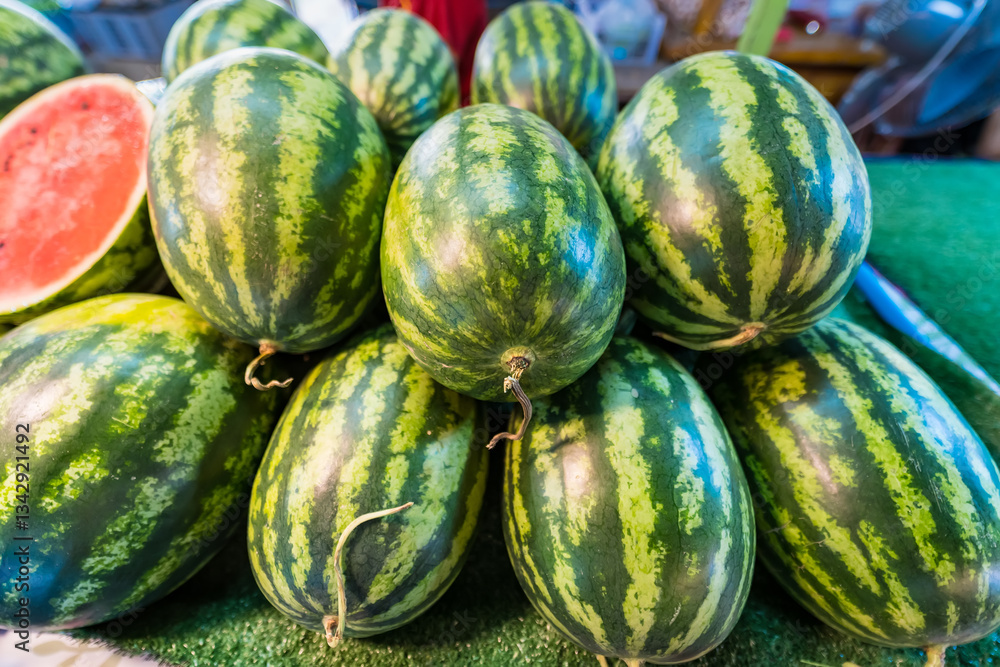 A pile of fresh whole watermelons with one cut open to reveal vibrant red flesh and dark seeds, displayed at a market stall A pile of fresh whole watermelons with one cut open to reveal vibrant red flesh and dark seeds, displayed at a market stall