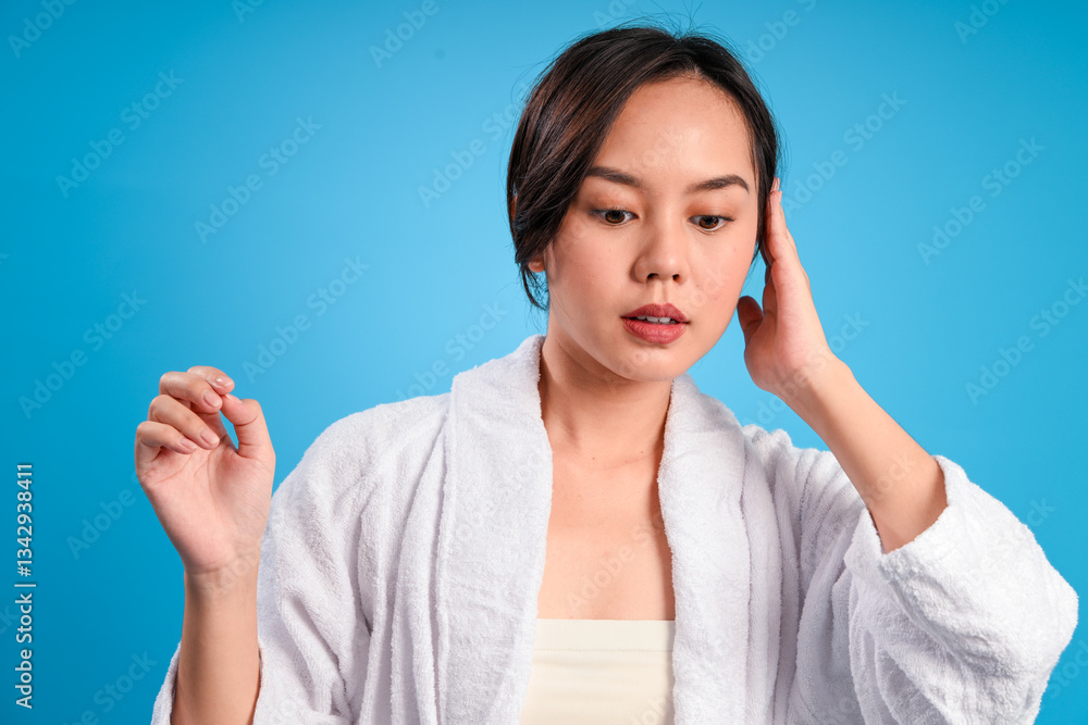 Asian woman in white bathrobe touching her head with one hand while looking down, standing against blue background, expressing calmness, reflection, or skincare awareness in soft lighting