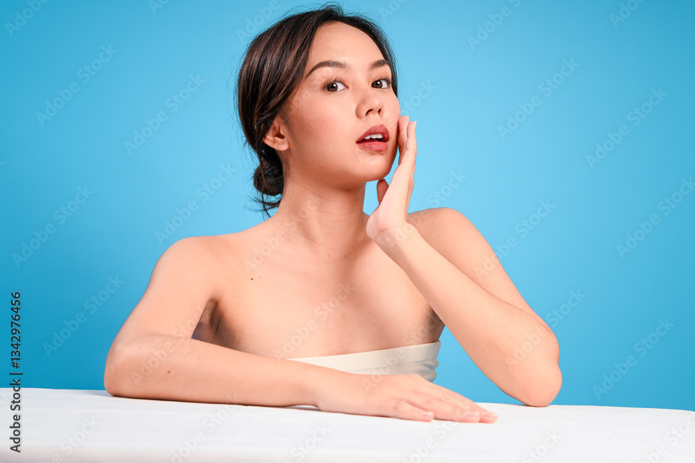 Asian woman with flawless skin and natural makeup posing topless against bright blue background, resting one arm on white table while gently pressing her cheek with one hand in calm expression Asian woman with flawless skin and natural makeup posing topless against bright blue background, resting one arm on white table while gently pressing her cheek with one hand in calm expression