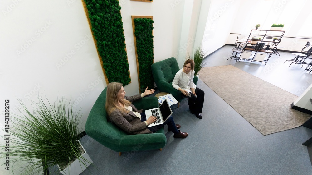 Fototapeta premium Two professionals collaborate in a modern office lounge with greenery and casual seating during a productive work session