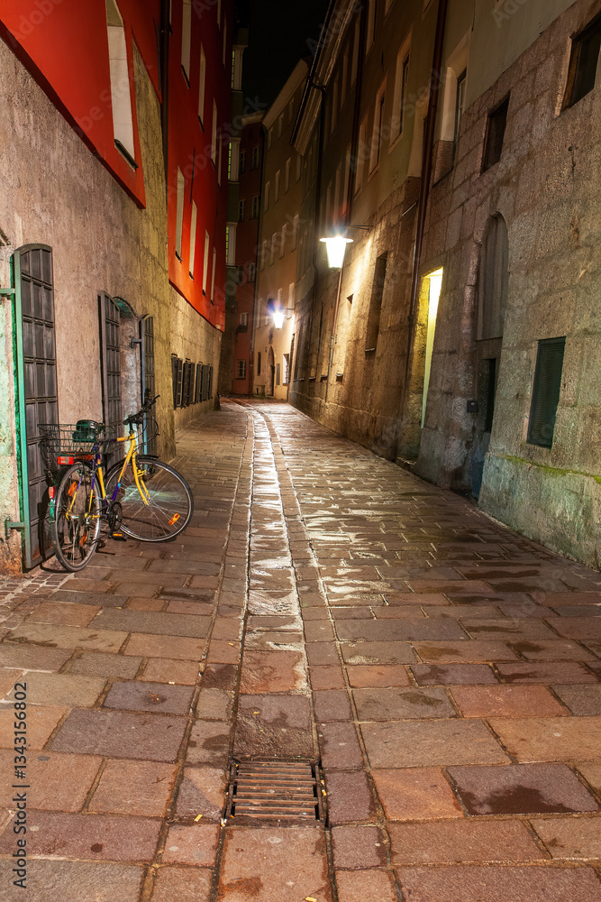 Fototapeta premium Quaint cobblestone alley in Innsbruck's historic center illuminated by warm streetlights. A colorful bicycle adds character to the scene, highlighting the city's blend of old and new