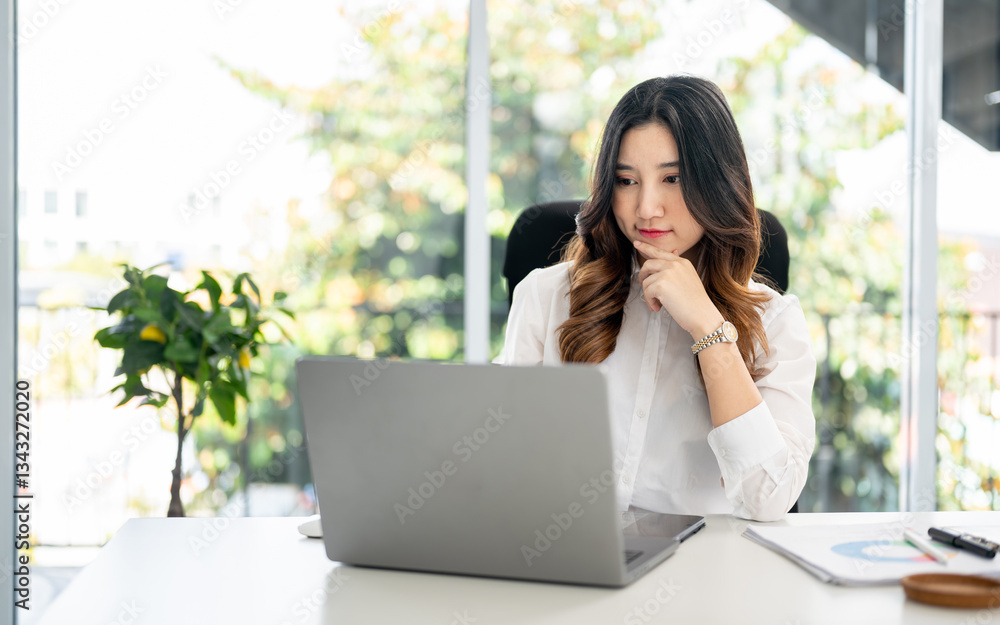 Businesswoman or female office worker putting a hand under chin, looking at laptop on working desk. Businesswoman or female office worker putting a hand under chin, looking at laptop on working desk.