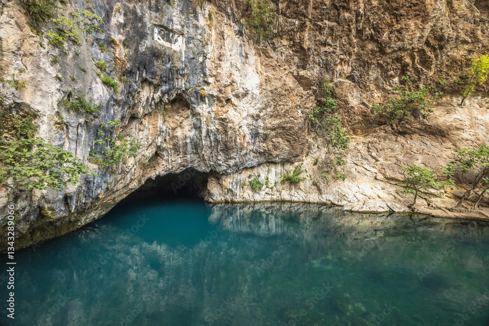 Fototapeta premium Buna river near the Blagaj Tekke monastery, Bosnia and Herzegovina, Europe. Cave opening at riverbank, with turquoise waters reflecting rock formations and vegetation in serene outdoor scenery.