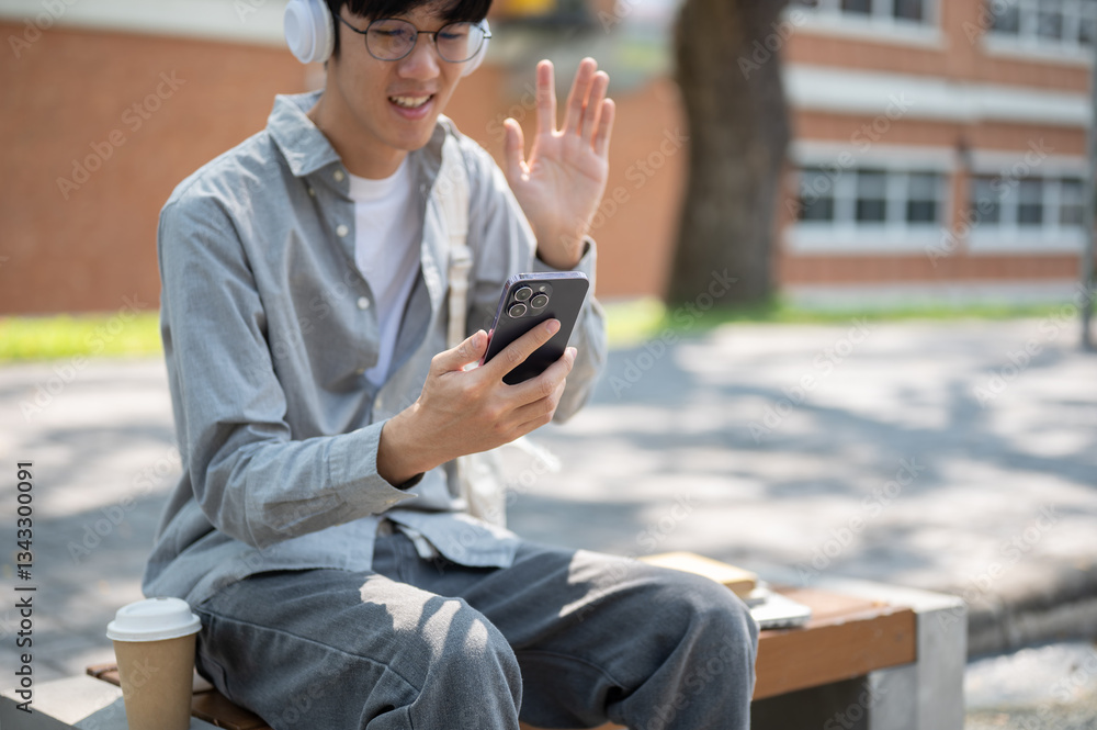 A smiling asian man with glasses is waving to his phone, making a video call or recording himself. A smiling asian man with glasses is waving to his phone, making a video call or recording himself.