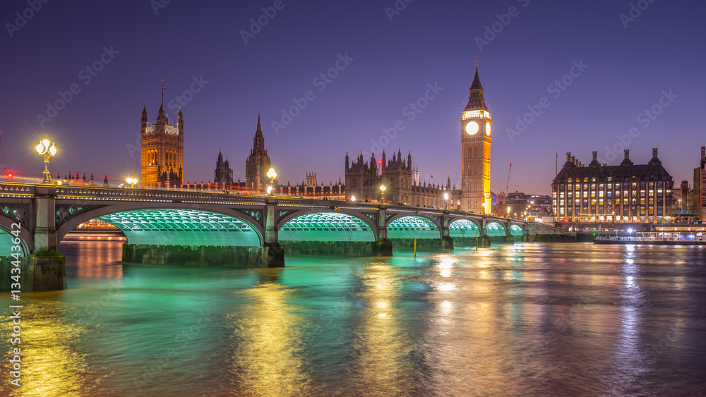 Naklejka premium The Houses of Parliament and Big Ben glow against the night sky, with Westminster Bridge elegantly lit. The tranquil river reflects the lights, showcasing London's iconic architecture.