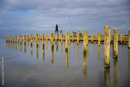 Holzpfähle in ruhigem Wasser mit bewölktem Himmel und entfernter Industriestruktur.