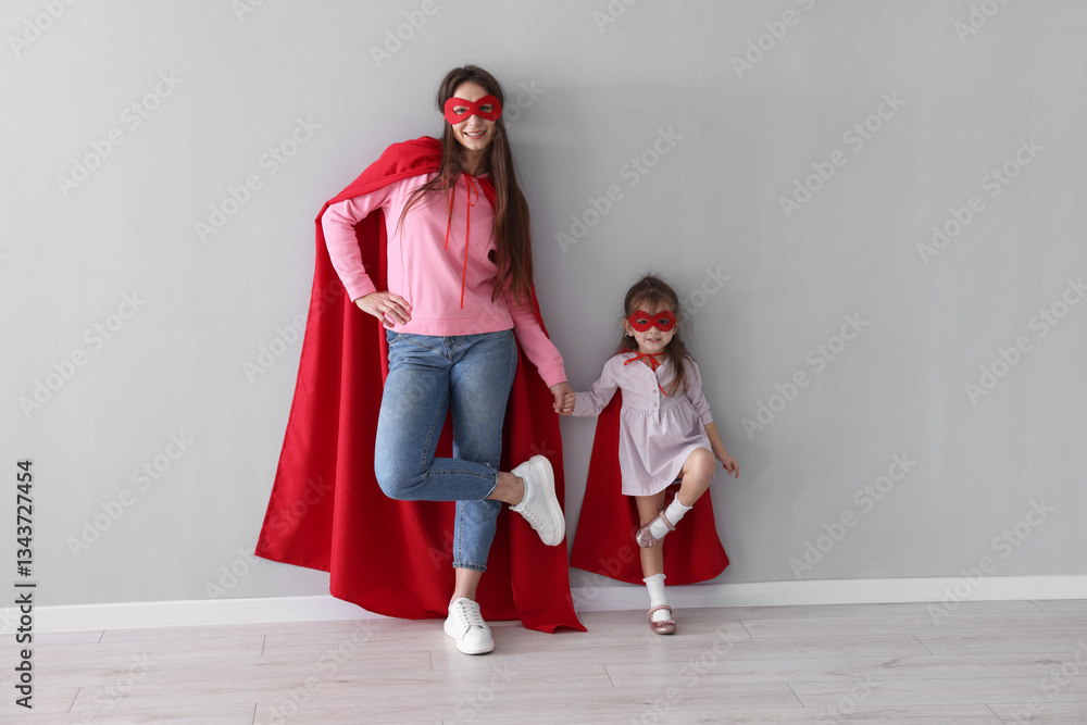 Fototapeta premium Mother and her cute little daughter wearing superhero costumes near gray wall indoors