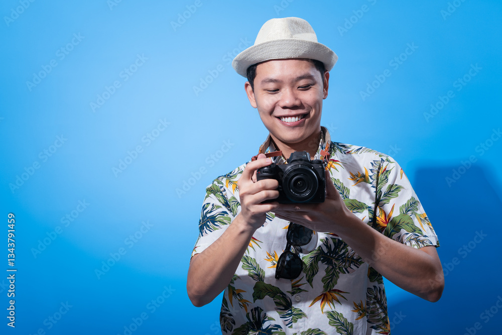 Obraz premium Asian man in floral shirt and white hat smiling while holding a camera and taking a photo, sunglasses hanging from neck strap, standing in front of bright blue background in studio setup