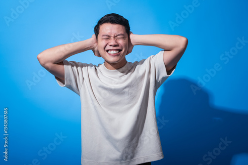 Asian man in a plain white t-shirt smiling with eyes closed and hands covering both ears, expressing joy, playfulness, or blocking noise, standing against a solid bright blue background