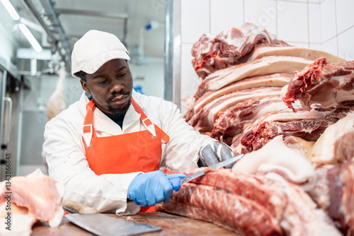 Professional butcher with sharp cleaver working with raw meat in slaughterhouse.