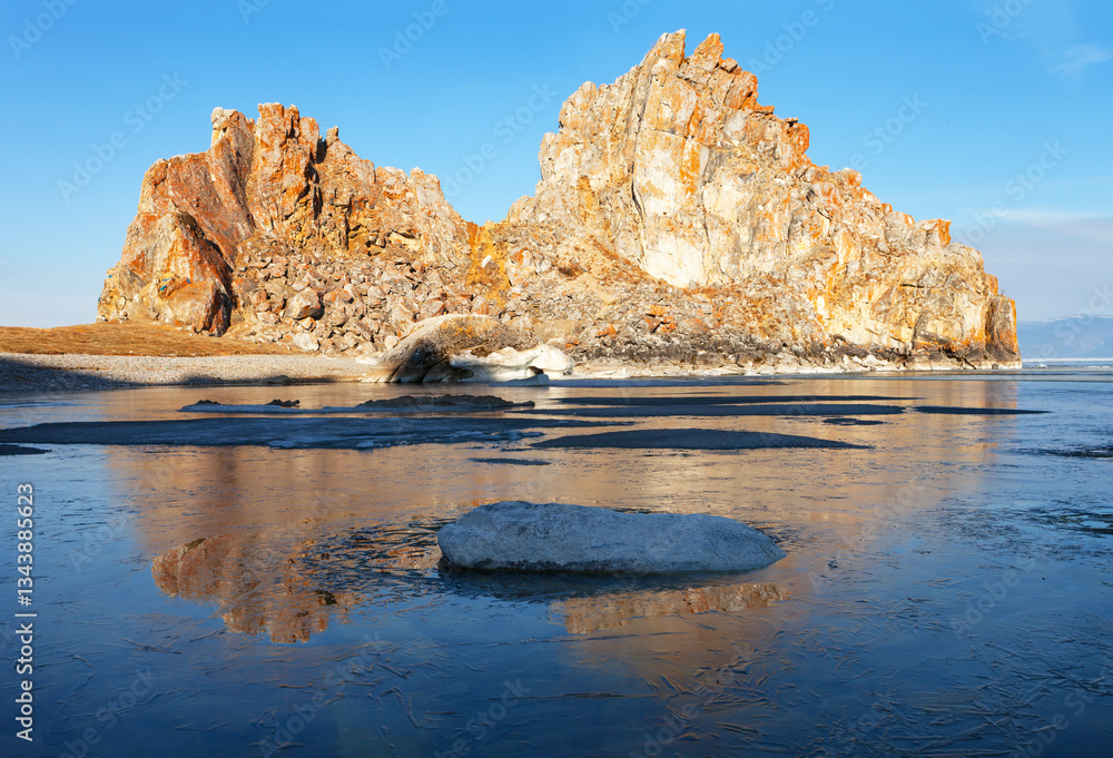 Obraz premium Spring morning on Baikal Lake. View of the famous Shamanka rock - a natural landmark of Olkhon Island in the rays of the rising sun. The bay is covered with a thin crust of blue ice. Spring landscape