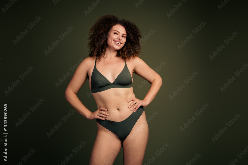 Obraz premium Confident young woman in green sportswear showcasing body positivity and natural beauty against a minimalist khaki background.