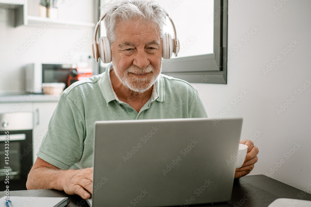 Smiling active modern white haired senior man with headphones sitting at home table using laptop in online connection surfing the net, following online course enjoying a coffee cup Smiling active modern white haired senior man with headphones sitting at home table using laptop in online connection surfing the net, following online course enjoying a coffee cup