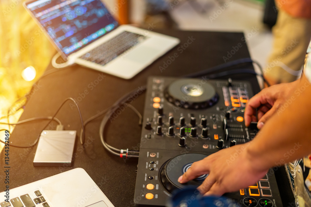 Obraz premium DJ at Work: The hands of a skilled DJ are expertly manipulating the controls of a mixing console, crafting music and rhythms for an unseen audience, with a laptop nearby and a hard drive ready.