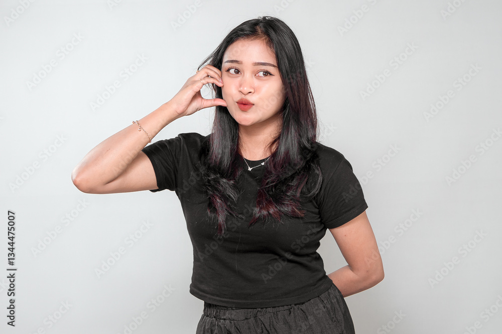 A smiling Indonesian woman with long black hair gestures a phone call sign with her hand. She wears a black t-shirt, expressing friendliness, approachability, and communication in a studio shot. A smiling Indonesian woman with long black hair gestures a phone call sign with her hand. She wears a black t-shirt, expressing friendliness, approachability, and communication in a studio shot.