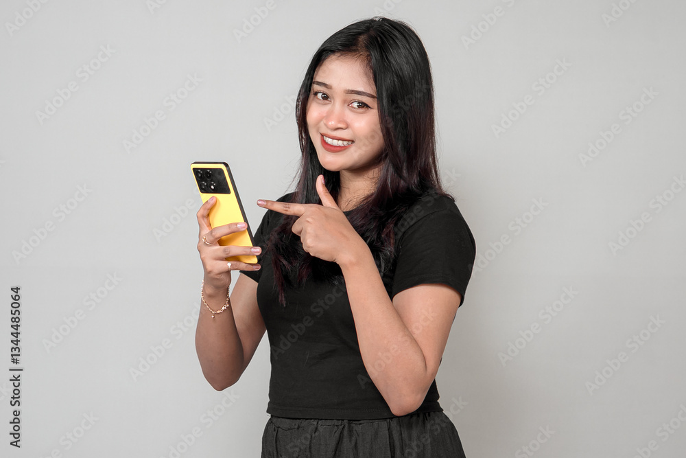 An Indonesian woman in a black outfit smiles while pointing at her yellow smartphone. She looks happy and confident, engaging with technology in a modern and stylish way. An Indonesian woman in a black outfit smiles while pointing at her yellow smartphone. She looks happy and confident, engaging with technology in a modern and stylish way.