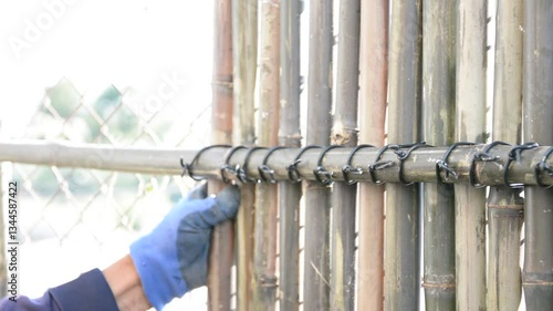 Worker install bamboo wall panels tied together with aluminum wire to cover a chain link fence.