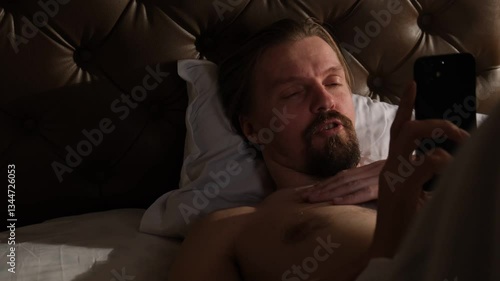 Young sexy handsome guy uses mobile phone before sleep lying in bed and chatting with women to meet and seduce. Attractive man with the help of smartphone meets mistress and flirts