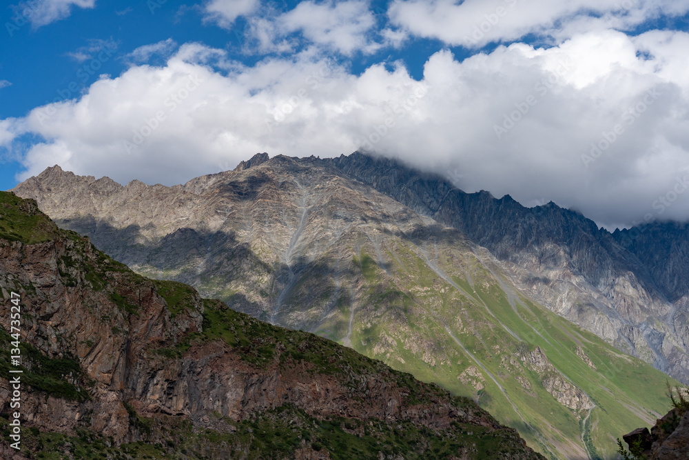 Fototapeta premium Rugged mountainous landscape with rocky peaks, lush green slopes, and scattered clouds casting shadows. A serene natural scene, ideal for nature, exploration, and outdoor themes.