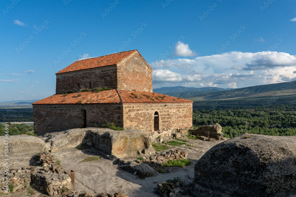 Ancient stone building with a red-tiled roof on rocky terrain, surrounded by ruins and a picturesque landscape of green fields, forests, and distant mountains under a blue sky. Ancient stone building with a red-tiled roof on rocky terrain, surrounded by ruins and a picturesque landscape of green fields, forests, and distant mountains under a blue sky.
