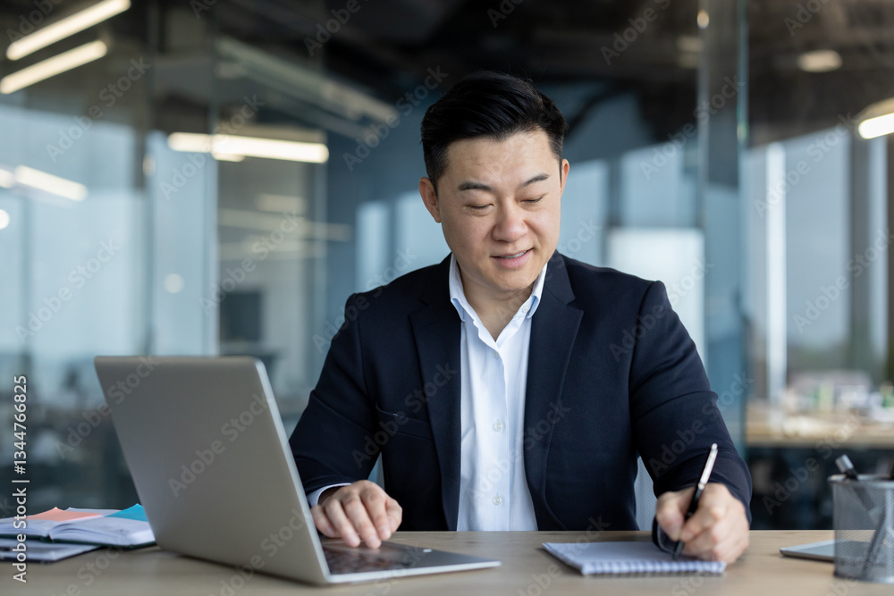 Smiling and successful young Asian male businessman sitting in the office at his desk, working on a laptop, making notes in a notebook Smiling and successful young Asian male businessman sitting in the office at his desk, working on a laptop, making notes in a notebook
