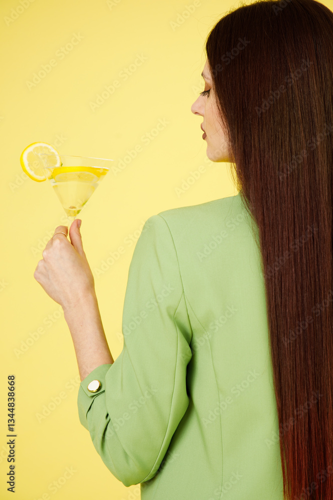 Fashion portrait of beautiful model in green suit with glass of lemonade against yellow background