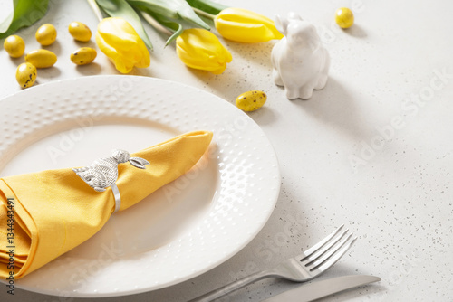Easter table setting with yellow tulips, white plates, bunny, eggs on white background. Happy Easter celebration. Copy space. Close up.