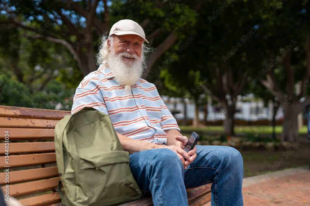 Fototapeta premium Elderly man with a long white beard and mustache, wearing a beige cap, striped short-sleeve sweater, and blue jeans, sitting on a wooden bench in a park, smiling while holding a smartphone 