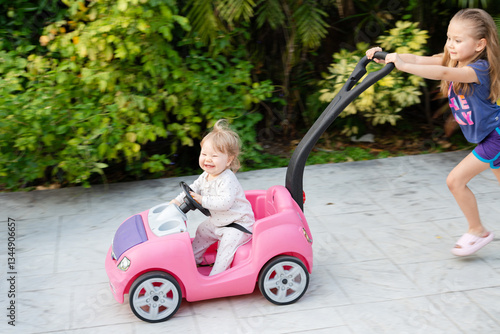 Happy baby in a pink toy car smiles while gripping the steering wheel, as an older sister joyfully pushes from behind capturing sibling fun and outdoor playtime. Authentic, candid shots