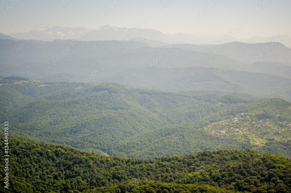 Fototapeta premium Expansive view of lush green forests and majestic mountains under hazy sky. Distant peaks blend into horizon, creating layers of tranquil scenery. Soft, natural lighting enhances serene atmosphere