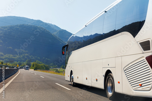 Scenic back view big luxury white comfortable tourist bus driving through highway on bright sunny day mountain landscape background. Travel and coach tourism concept. Trip and journey by vehicle
