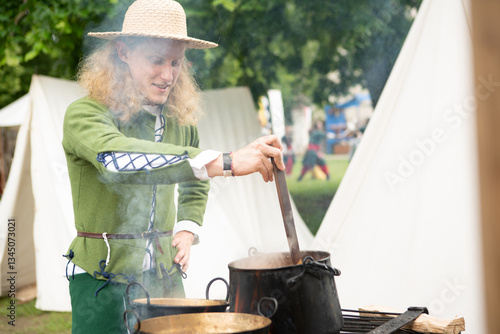 Obraz na plátně Smiling young Man in medieval attire straw hat and green tunic, cooks over open flame at historical reenactment event