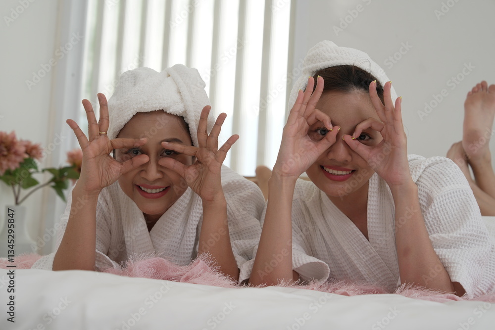 Asian mother and daughter in bathrobes laying on bed in bedroom, covering their eyes with their hands, having fun on a relaxing day, clean skin.