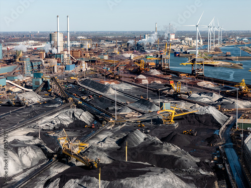 Steel industry in IJmuiden, Netherlands, showing coal storage, conveyors, and emissions. Wind turbines stand in contrast to fossil fuel-based production and industrial pollution. Aerial view.