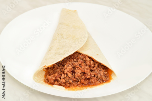Tortilla wrap with stewed mixed pork and beef minced meat with peeled and chopped tomatoes in tomato juice on the white plate on the light gray marble table texture. Close-up. Selective focus.