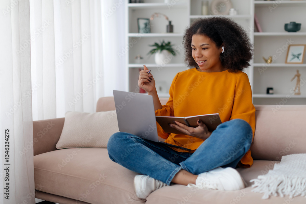 Young happy black woman in headphones multitasking at home, writing in notebook while using laptop, happy african american female attending online course or watching webinar, sitting on couch Young happy black woman in headphones multitasking at home, writing in notebook while using laptop, happy african american female attending online course or watching webinar, sitting on couch