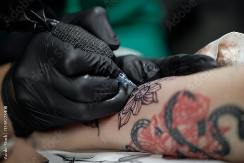 Close up tattoo master professional tattoo artist in black gloves making a rose tattoo on a man's forearm. He is holding a tattoo machine.