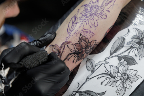 Close up tattoo master professional tattoo artist in black gloves making a rose tattoo on a man's forearm. He is holding a tattoo machine.