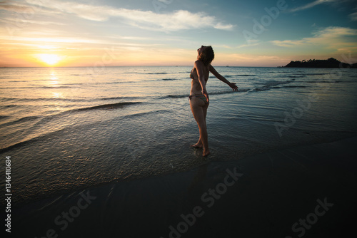Obraz na płótnie A woman in a swimsuit greets the sunrise on the beach, embodying relaxation and tranquility in a serene morning setting. w salonie