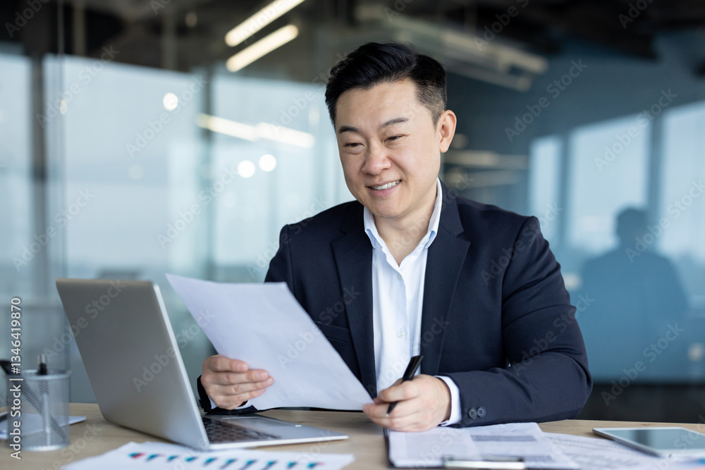 Smiling young Asian man in a suit working in the office at a desk with a laptop and reading documents and an agreement he is holding in his hands Smiling young Asian man in a suit working in the office at a desk with a laptop and reading documents and an agreement he is holding in his hands