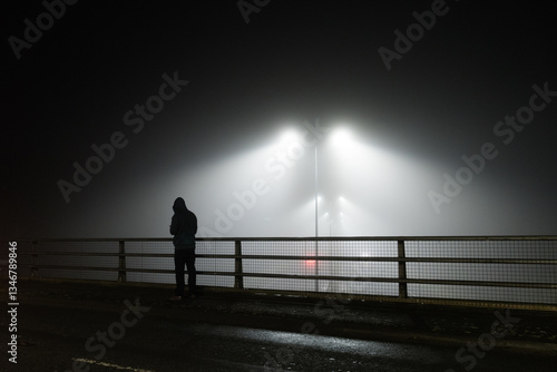 A mysterious unknown hooded figure standing on a bridge over a motoway, silhouetted by street lights on a foggy eerie night