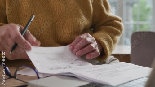 Senior elegant woman sits at table viewing papers and utility bills. Aged woman works remotely using laptop computer. Older focused businesswoman making notes. Female elegant writer creative process
