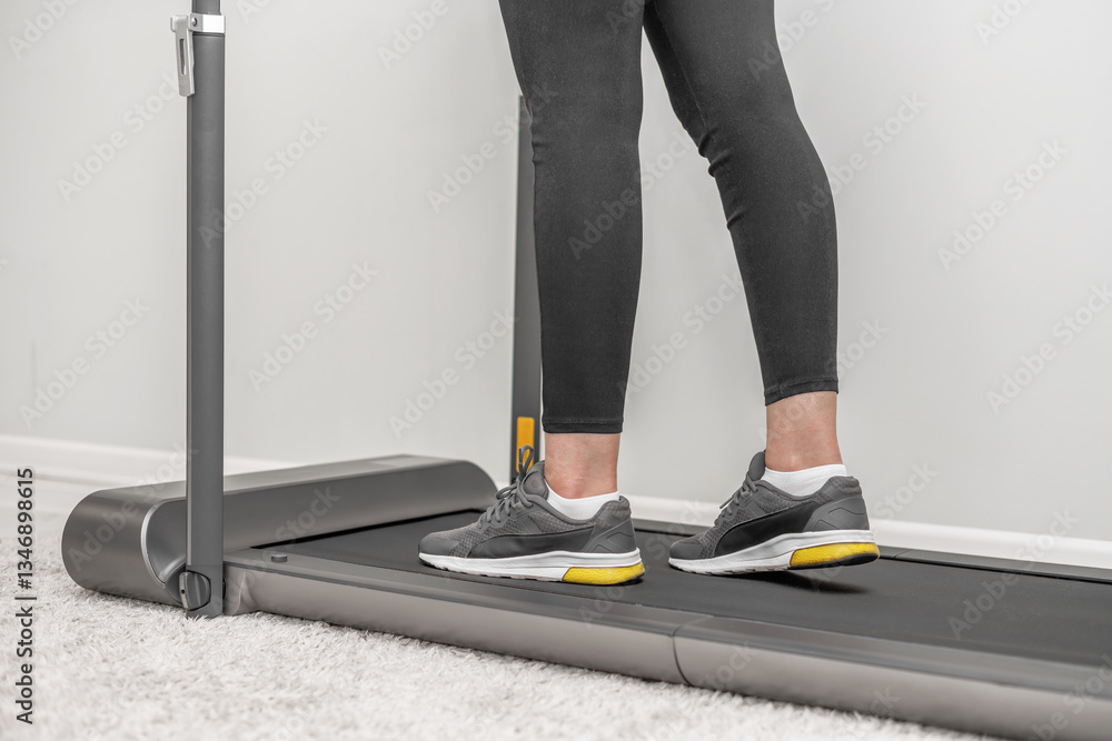 A person wearing black leggings and grey sneakers, with yellow soles is walking on a modern treadmill placed on a soft carpet in a minimalistic home setting.
