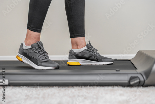A person wearing black leggings and grey sneakers, with yellow soles is walking on a modern treadmill placed on a soft carpet in a minimalistic home setting.